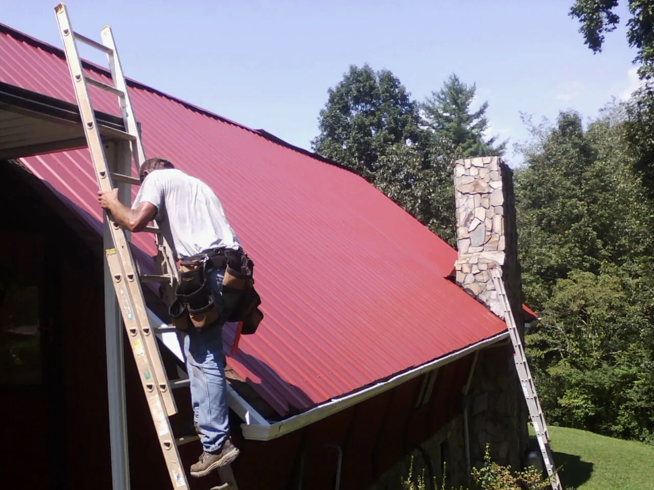Roofer on a ladder beside a red metal roof and stone chimney — Soffit & Fascia Repair by Roofing Brothers in Coward